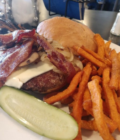 A juicy burger topped with bacon, onions, and mayo, served with sweet potato fries and a pickle slice.
