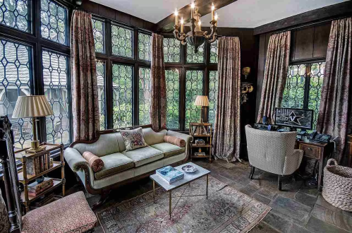 Cozy room with large windows, a vintage sofa, a desk, and warm lighting, surrounded by greenery outside.