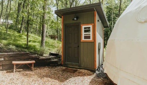 A small, modern cabin with a dark door and orange trim, surrounded by trees and a wooden bench nearby.
