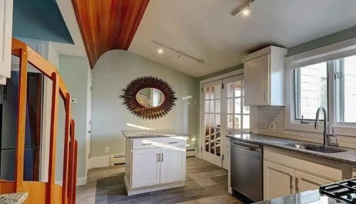 Bright kitchen with modern appliances, a central island, and a unique wooden ceiling design. Sunlight streams through windows.