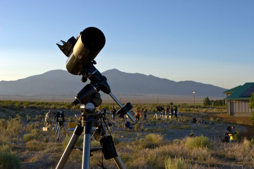 A telescope in the foreground with a mountain backdrop, and people setting up equipment in the distance during sunset.
