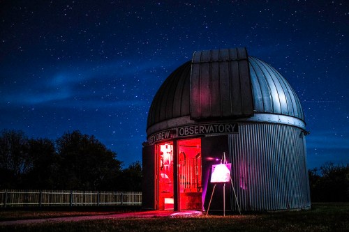 A dome-shaped observatory under a starry night sky, illuminated by red light, with a sign outside.