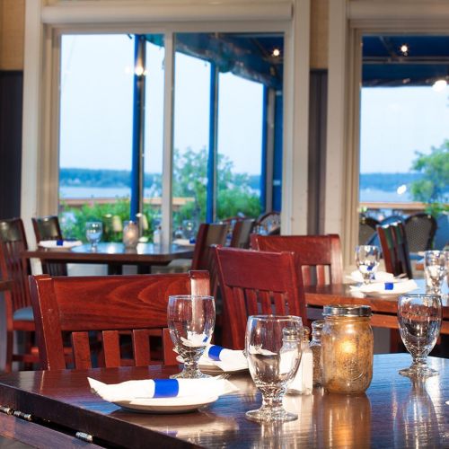 Cozy restaurant interior with wooden tables, glassware, and a view of the outdoors through large windows.