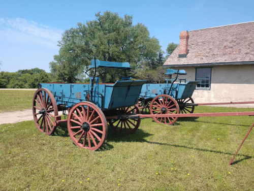 Two vintage blue wagons with red wheels parked on green grass near a building under a clear blue sky.