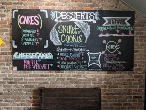 Chalkboard menu listing desserts: cakes, cheesecakes, pies, and kids' options with prices. Rustic brick background.