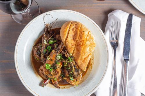 A bowl of shrimp in sauce with green onions, served with a piece of bread, alongside a glass of red wine and cutlery.
