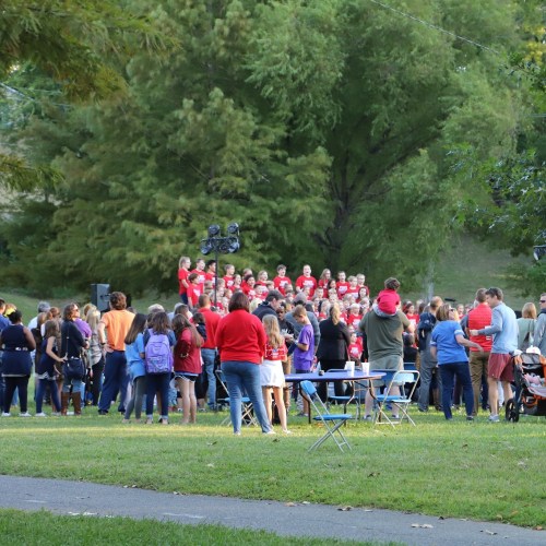 A large group of people gathers outdoors, watching a choir performance in front of a backdrop of trees.