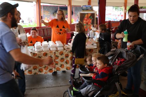A busy market scene with people in pumpkin-themed shirts, children in strollers, and a festive atmosphere.