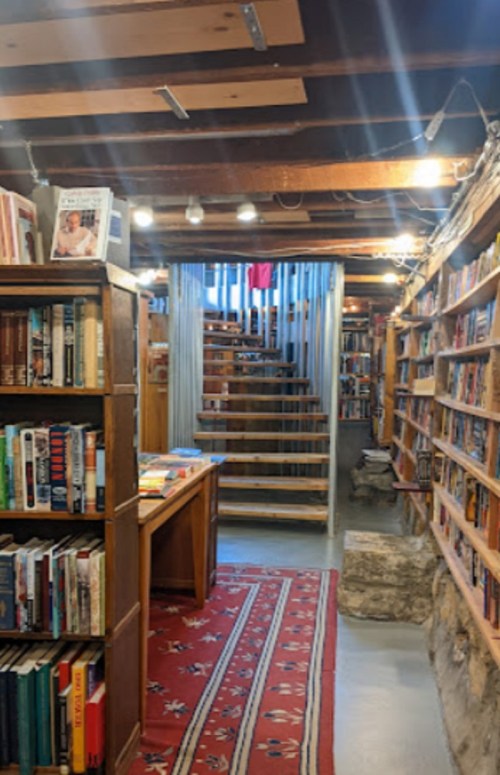 A cozy bookstore interior with wooden shelves, a red rug, and a staircase leading to an upper level.