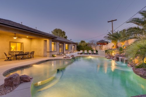 A serene backyard pool with a waterfall, surrounded by palm trees and lounge chairs, at dusk.