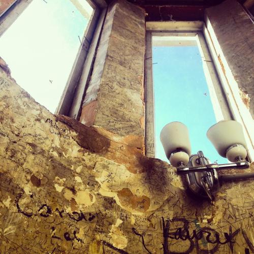 Interior of a room with peeling walls covered in graffiti, featuring two windows and a light fixture.