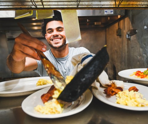 A smiling chef serves food onto plates in a busy kitchen, showcasing a dish with eggs and bacon.