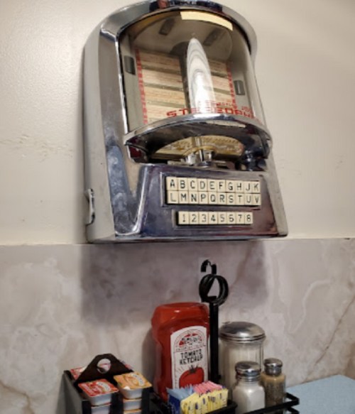 A vintage jukebox mounted on a wall, with a condiment holder featuring ketchup, mustard, and sugar packets below.
