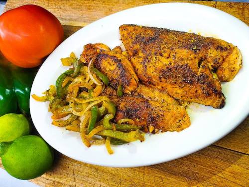 A plate of seasoned fish fillets with sautéed peppers and onions, accompanied by fresh tomatoes and limes.