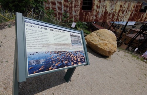 Sign about a giant fossil thrombolite next to a large rock, with a rusted building in the background.