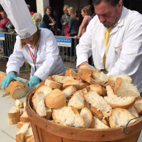 A chef and a man in a white coat prepare large amounts of bread in a wooden barrel at a busy outdoor event.