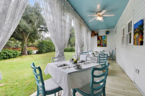A charming outdoor dining area with white tables, blue chairs, and sheer curtains, surrounded by greenery.