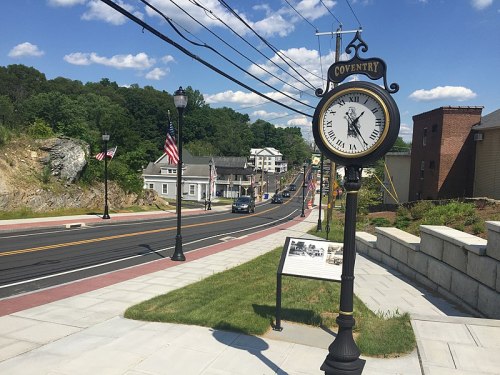 A street view of Coventry with a vintage clock, American flags, and buildings under a blue sky with clouds.