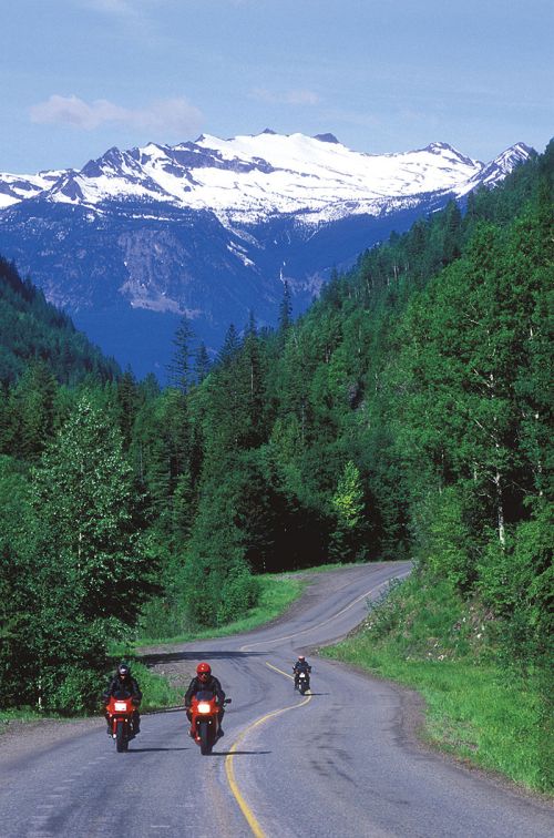 Two motorcyclists ride along a winding road surrounded by lush greenery and snow-capped mountains in the background.