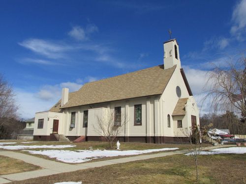 A white church with a brown roof and steeple, surrounded by grass and patches of snow under a blue sky.
