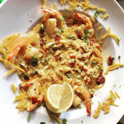 A bowl of shrimp topped with melted cheese, green onions, and a lemon wedge, garnished with shredded cheese.