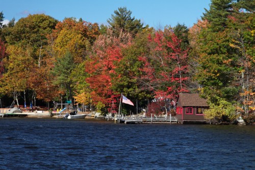 A serene lake scene with vibrant autumn trees, a red cabin, and an American flag by the water's edge.