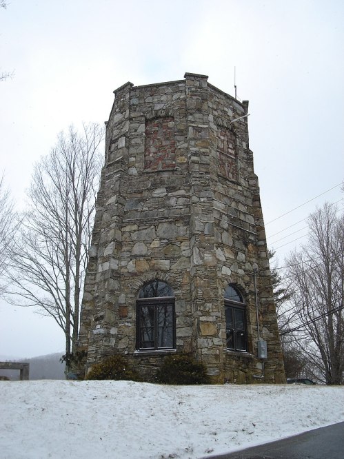 A tall, stone tower with arched windows, surrounded by snow and bare trees under a cloudy sky.