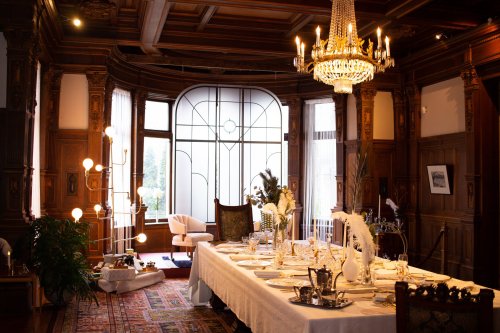 Elegant dining room with a long table set for a meal, featuring ornate woodwork, a chandelier, and decorative plants.