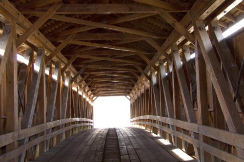 Interior view of a wooden covered bridge, featuring a curved wooden floor and beams, leading to a bright exit.