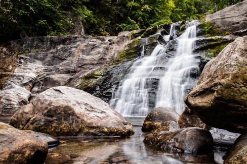 A serene waterfall cascades over rocky terrain, surrounded by lush greenery and smooth stones in a tranquil setting.