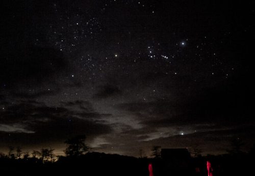 Two silhouetted figures stand under a starry night sky, with bright stars and clouds visible.