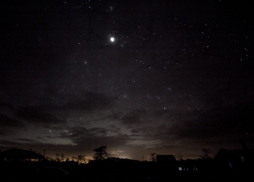 A starry night sky with scattered clouds and a bright planet visible, silhouetted trees and distant lights on the horizon.