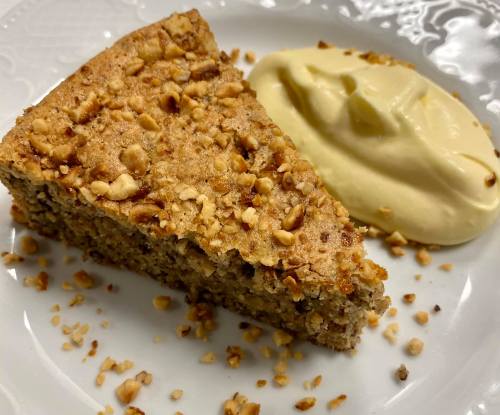 A slice of nutty cake topped with crushed nuts, served with a dollop of creamy custard on a white plate.