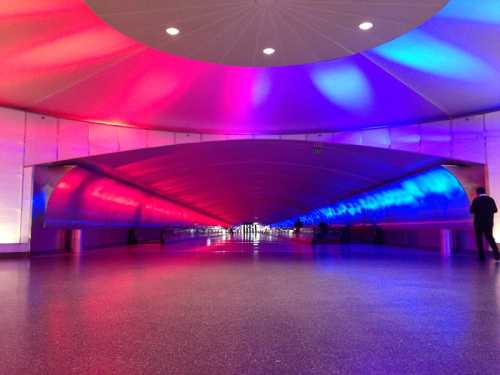 A modern, brightly lit tunnel with colorful red and blue lighting, featuring smooth, curved walls and a polished floor.