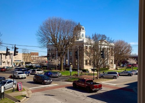 A busy intersection with cars, featuring a historic courthouse and trees under a clear blue sky.