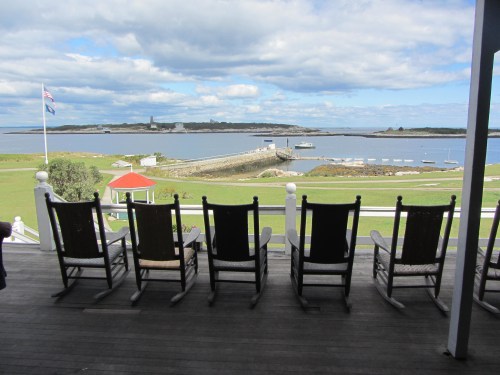 View from a porch with rocking chairs overlooking a calm harbor and distant lighthouse under a cloudy sky.