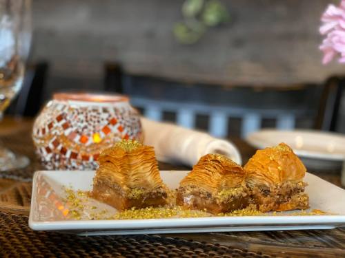 A plate of baklava topped with crushed pistachios, set on a table with a decorative candle in the background.
