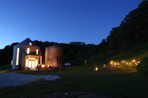 A modern building with silos at dusk, surrounded by a grassy area and lit by string lights and a campfire.