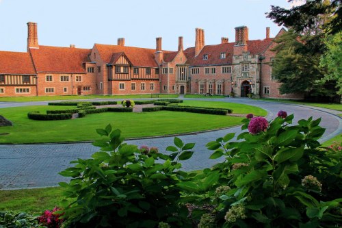 A grand brick mansion with a manicured lawn and flower beds, surrounded by trees and a circular driveway.