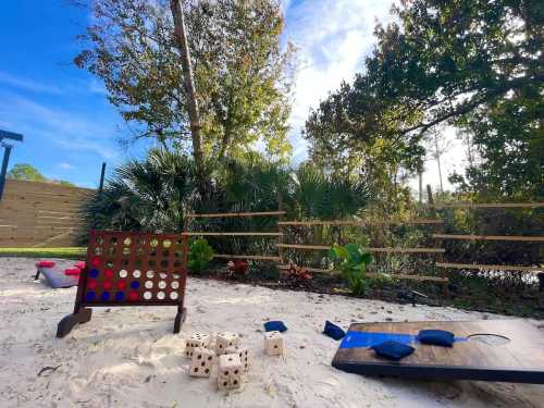 Outdoor game area featuring a Connect Four set, cornhole boards, and dice on sandy ground with greenery in the background.