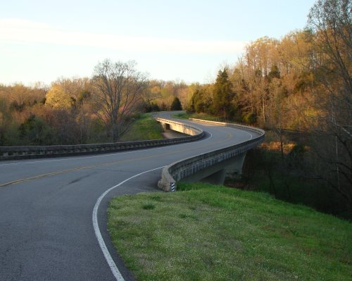 A winding road curves over a bridge, surrounded by trees and greenery under a clear sky.