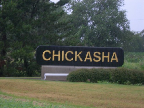 Sign for Chickasha, Oklahoma, with bold yellow letters on a black background, surrounded by greenery.