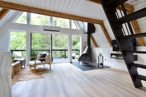 A modern, airy living space with large windows, wooden beams, and a sleek fireplace, surrounded by greenery.