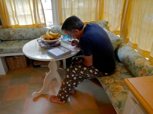 A person in pajamas sits at a table, writing in a notebook, surrounded by a cozy kitchen with yellow curtains.
