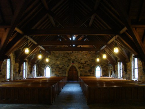 Interior of a rustic church with wooden beams, stone walls, and rows of wooden pews, illuminated by hanging lights.