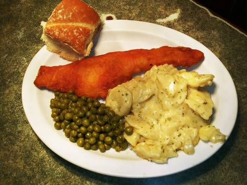 A plate with fried fish, creamy pasta, green peas, and a roll on the side.