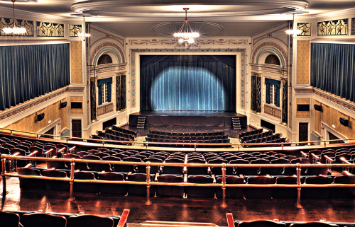 A grand theater interior with empty seats, a stage draped in blue curtains, and ornate architectural details.