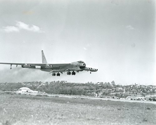 A large military aircraft is landing over a suburban area, with buildings and trees in the background.