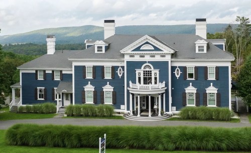 A large blue mansion with white trim, featuring a grand entrance and surrounded by lush green grass and mountains in the background.