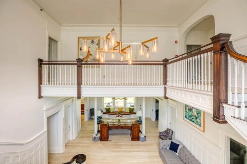 A spacious, bright foyer with a modern chandelier, wooden railing, and elegant decor, leading to a living area.